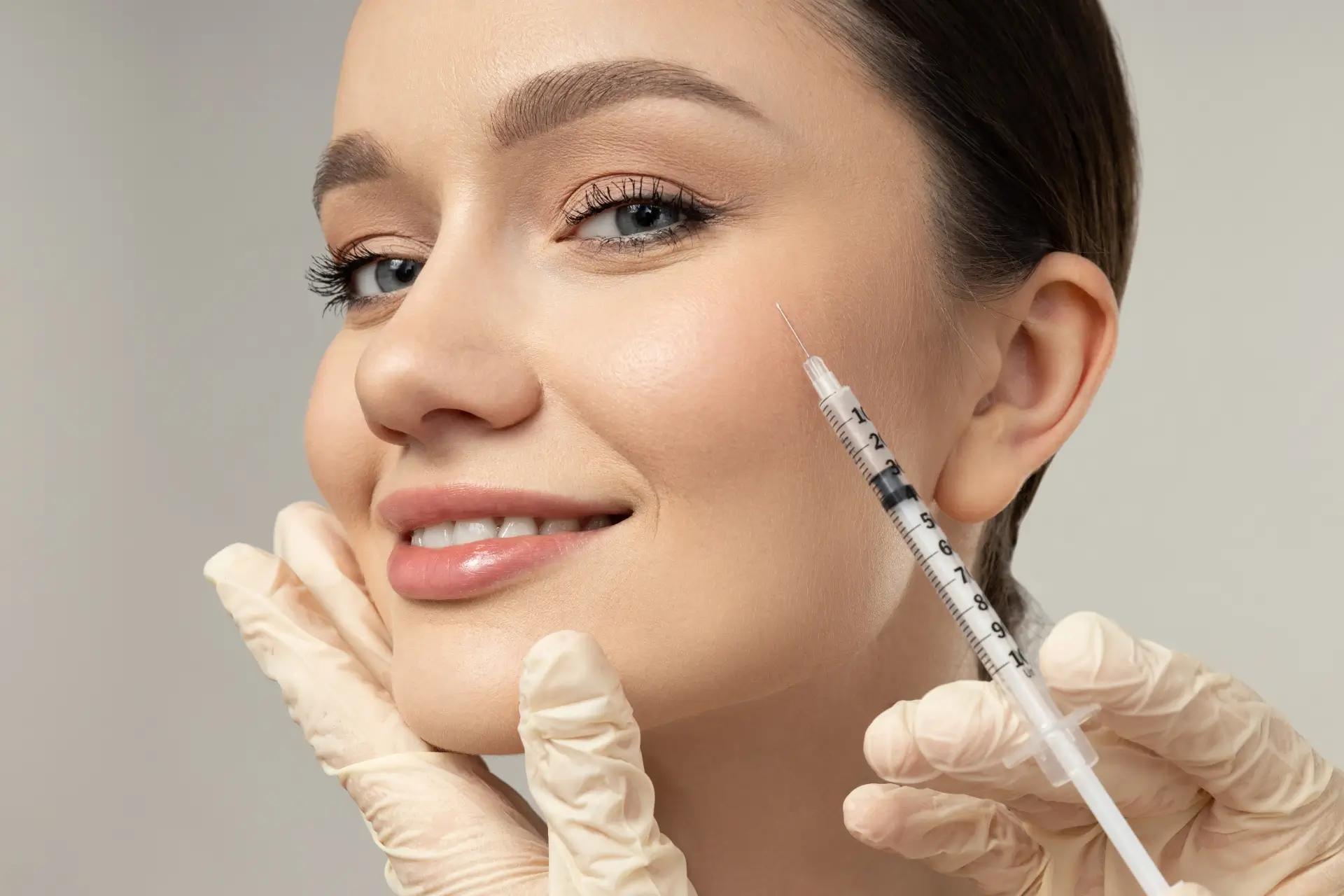 A smiling client rests her chin on her hand while a gloved practitioner holds a fine syringe beside her cheek