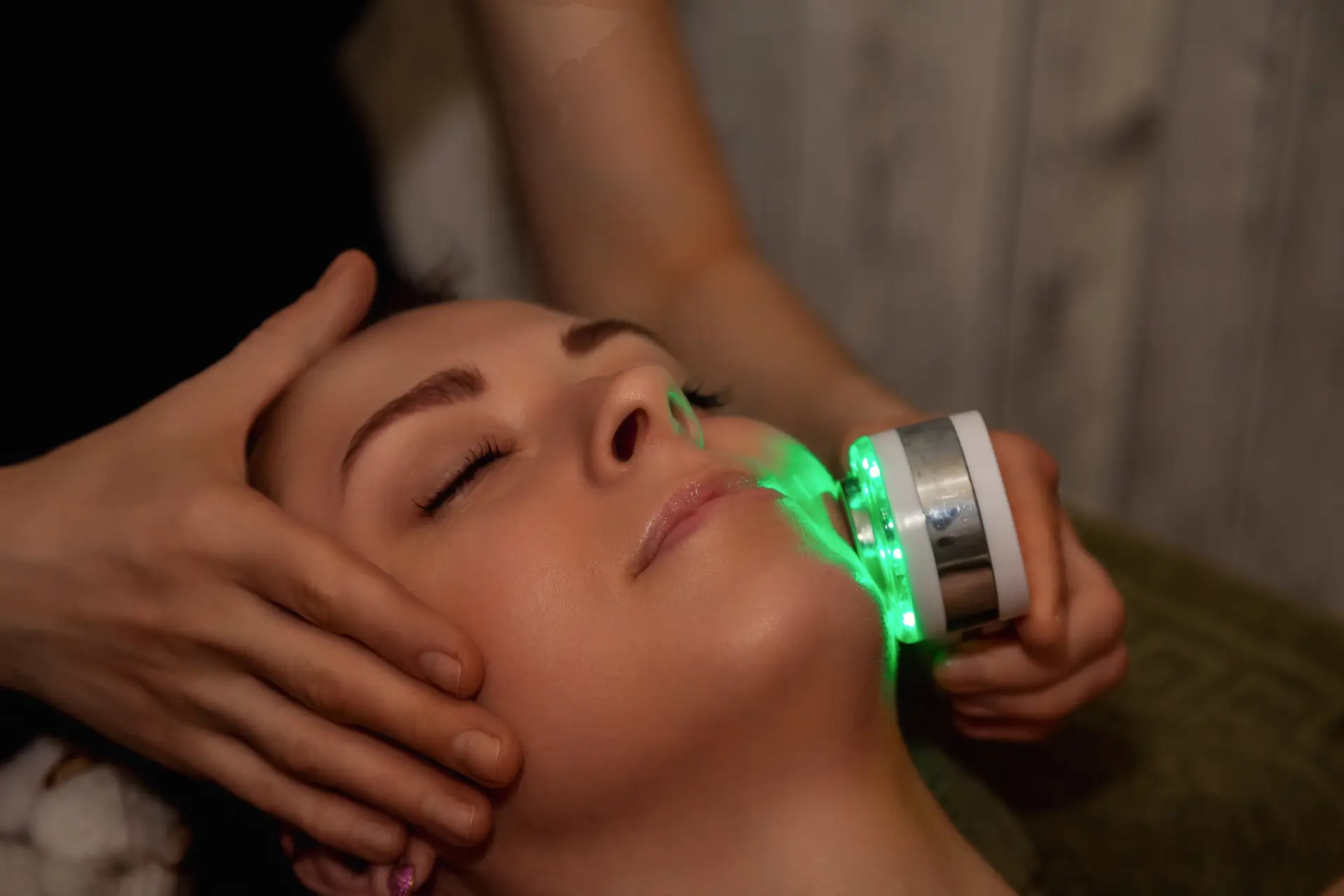 A handheld laser emits bright green light across a client's cheek while a practitioner steadies her head in a darkened treatment room