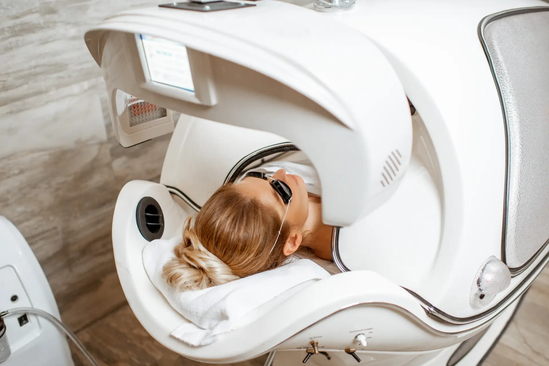 A client wearing protective eyewear reclines beneath a large medical laser device in a bright clinic treatment room