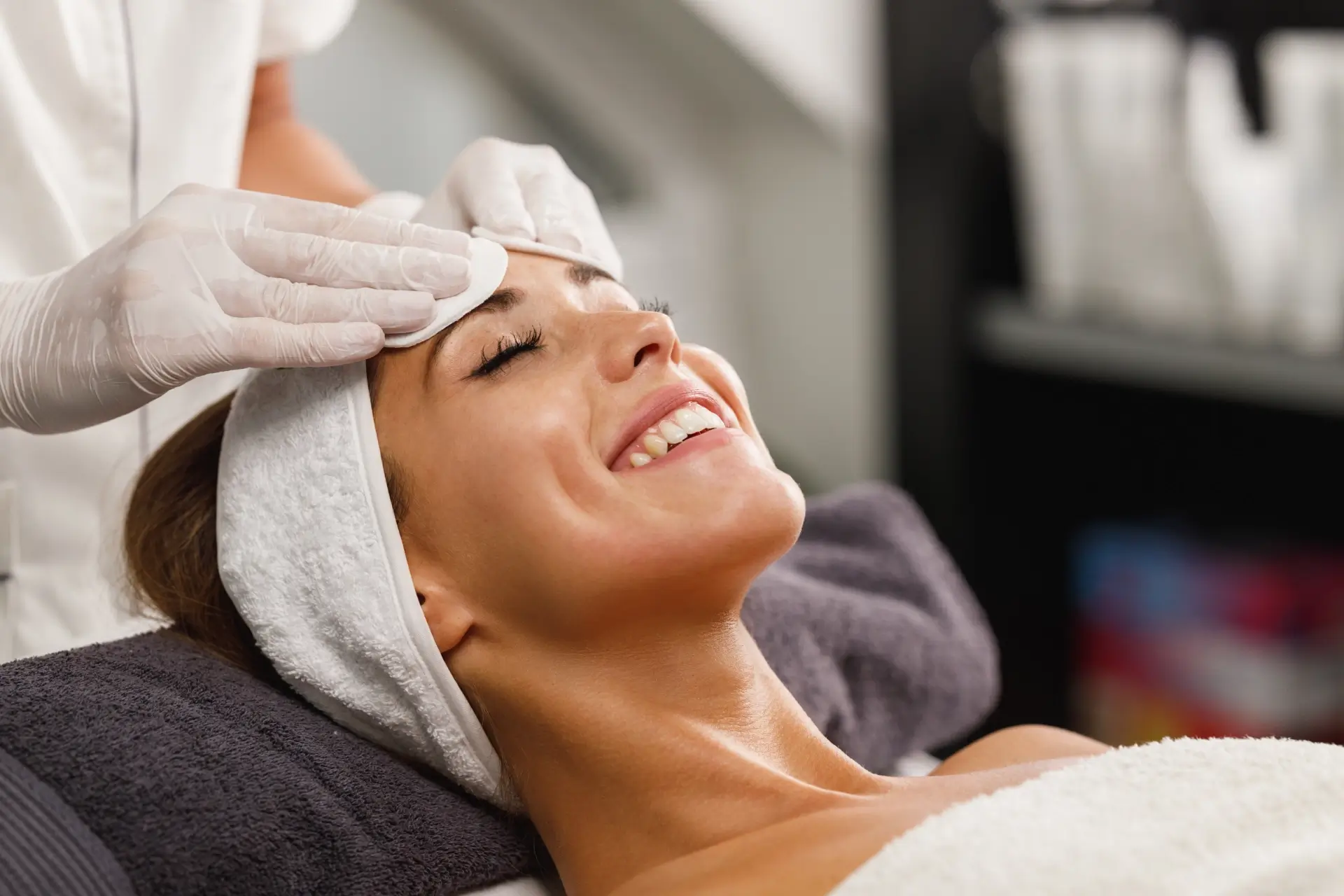 A smiling client rests on a treatment bed with a white headband while a practitioner's gloved hands support her forehead