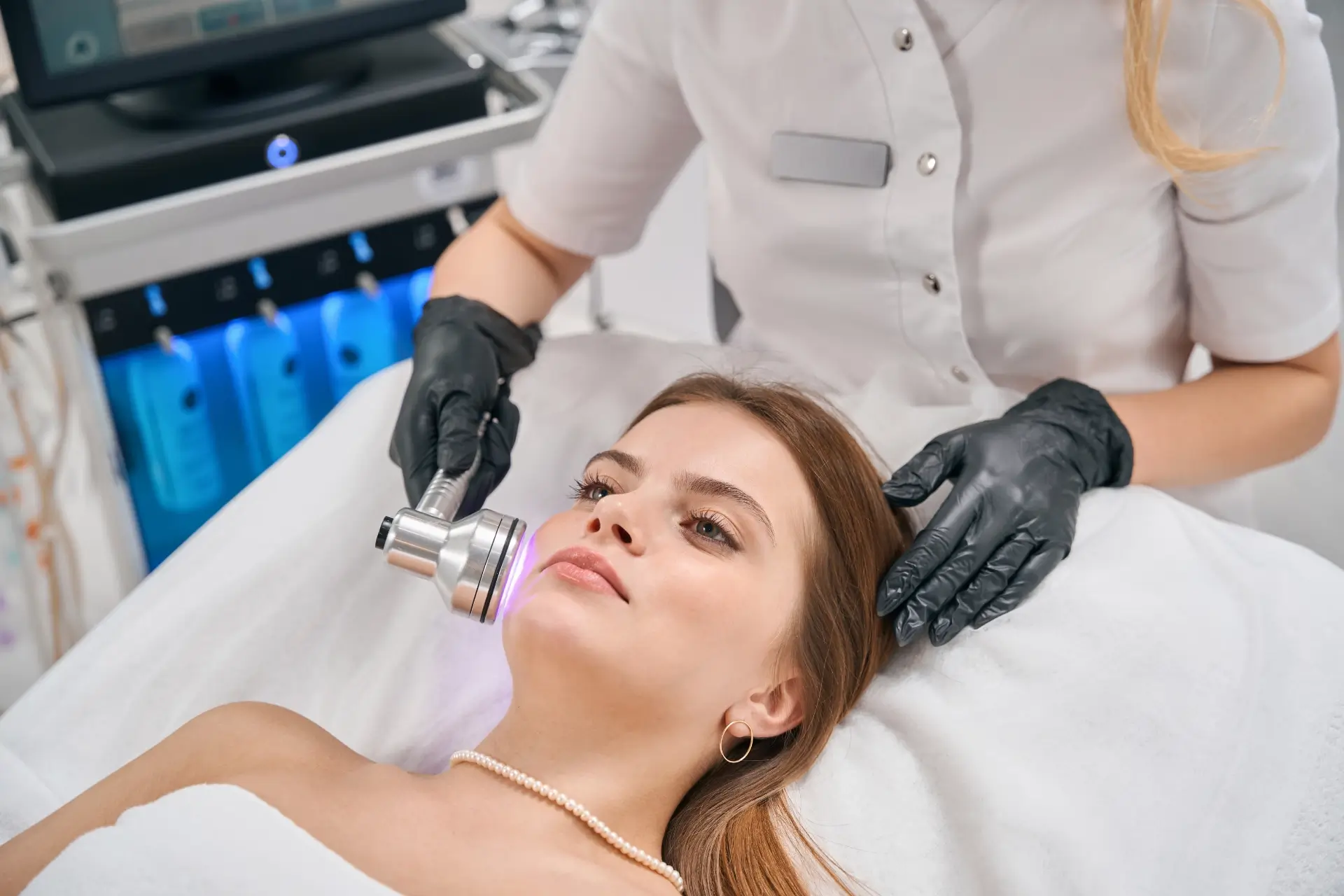 A practitioner in gloves guides a light-based handpiece across a reclining client's cheek during a laser skin rejuvenation session